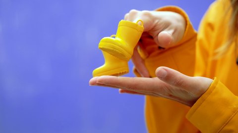 Cropped detail of woman holding a pair of rainy boots in blue background. Horizontal side view of hands playing funny steps with yellow galoshes isolated in blue background. Conceptual backgrounds.