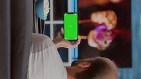 Static tripod shot of man holding smartphone with green screen watching online video content with girlfriend sitting on sofa. Couple enjoying social media videos on mobile phone with chroma key .