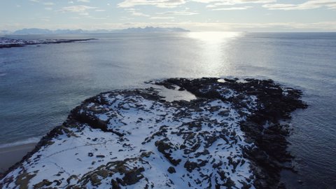 Truck left aerial shot of a snow covered rocky outcrop into the ocean and beach in Norway with bright glare off the ocean