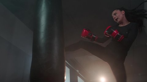 Strong, aggressive female fighter kicks a punching bag. Sporty angry woman boxing professionally in a dark gym. Slow motion. The concept of a strong independent woman, feminism. High quality 4k