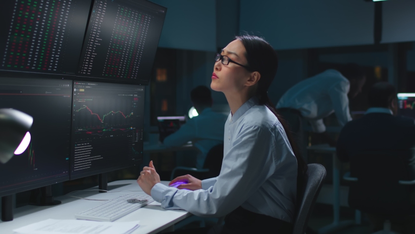Asian woman trader look at multi-screen workstation with graphs and data working late in dark office. 
