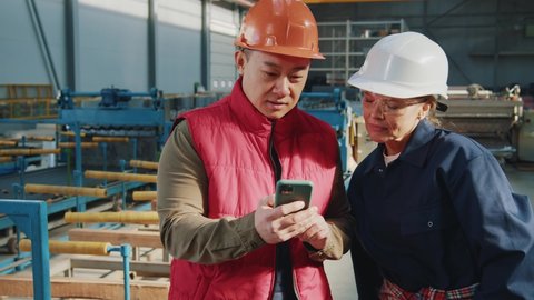 Portrait of two factory engineers technicians with helmet use mobile phone standing in factory. Woman and asian man. Team