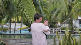 In the fresh air with beautiful green trees, a young father raises and lowers his small child with his hands, holding him tightly. The child is less than a year old. Slowmo video.