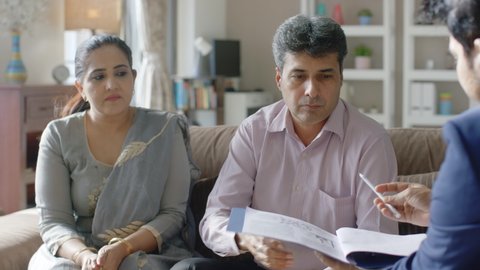 Upset Indian Man and Woman sitting together on a couch in an interior house setup signing divorce papers or. documents while ex-wife walks away in anger. concept of separation couple marital conflicts