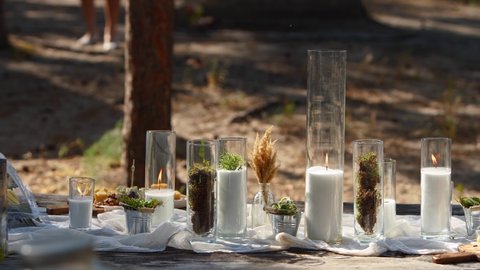 Wedding party banquet outdoors in forest. Dining table decorated in boho style with candles, white cloth, flowers, served with plates, tableware, dishes, meals, stemware and drinks. Holiday catering.