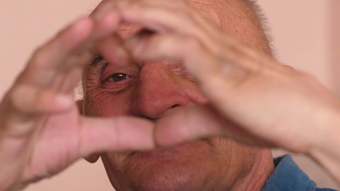 Portrait of a caucasian cheerful elderly man 70 years old showing a heart gesture with his hands. Pensioner congratulates on Valentine's Day. Elderly man in love heart gesture.Selective focus