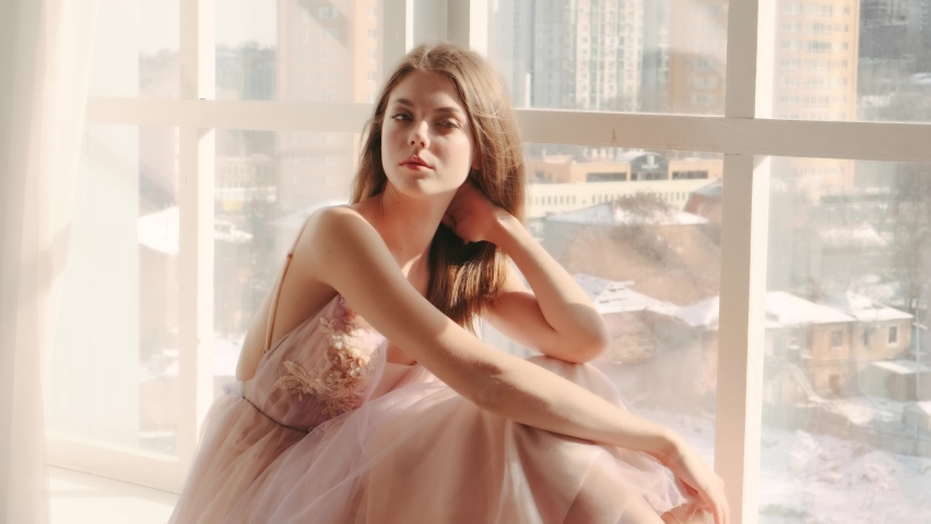 Portrait adult beautiful blonde girl princess sitting on windowsill. Young woman enjoys view from large panoramic window. Delicate makeup cute face gentle makeup, evening prom pink dress ball gown.