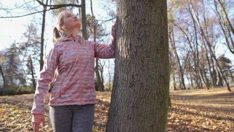 A satisfied blonde stands next to a large tree and admires it.