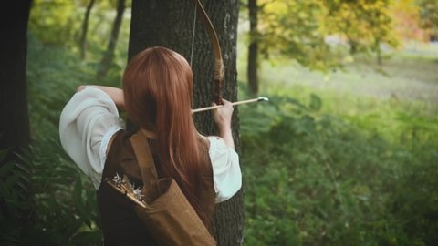 Video with noise. Woman Vintage archer hiding near tree, shooting an arrow from bow. Medieval hunter girl in summer forest Girl holding weapons in hands. Costume medieval warrior. Back, rear view
