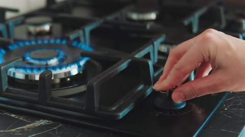 Person using kitchen burner with hand. Ignited bright blue flame, fire for cooking. Background black color cooktop, hob. Gas stove being turned on by lit burner. Natural gas inflammation, close up.