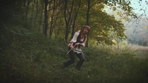 Adult fantasy woman archer walks in foggy forest. girl holds wooden bow in her hands. Lady medieval hunter. Vintage warrior archeress. clothes brown leather pants, white shirt jacket, bag with arrows