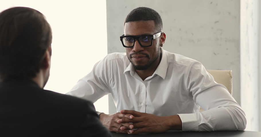 Smiling confident young Black man job applicant sit at desk at employer office talk to male recruiter answer questions on interview. Two diverse male business partners hold negotiations discuss deal Royalty-Free Stock Footage #1089340169