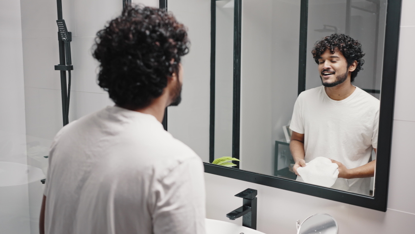 Young Asian Indian man wipes face with white terry towel after washing in hotel bathroom. Curly-haired guy looks in mirror in morning closeup Royalty-Free Stock Footage #1089342379