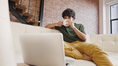 Hindu man watches comedy movie on laptop enjoying cup of hot tea. Young guy with curly hair sits on couch smiling and relaxing at home closeup