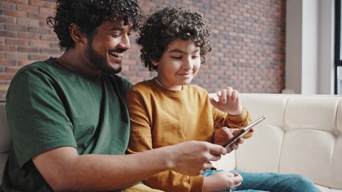 Father shows son interesting videos about animals on tablet. Smiling curly-haired man and schoolboy sit on sofa in apartment living room closeup