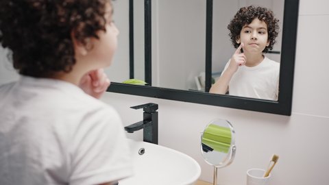 Cute biracial boy with curly hair puts moisturizing cream on cheeks. Child looks in mirror standing in bathroom. Skincare routine close view