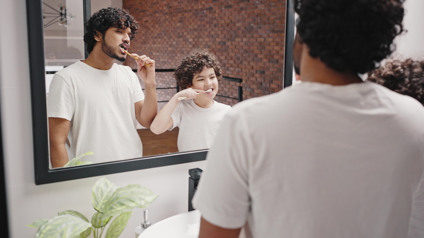 Asian Indian man and biracial brother brush teeth against mirror after waking up in morning. Curly-haired siblings keep routine in bathroom closeup Royalty-Free Stock Footage #1089342465