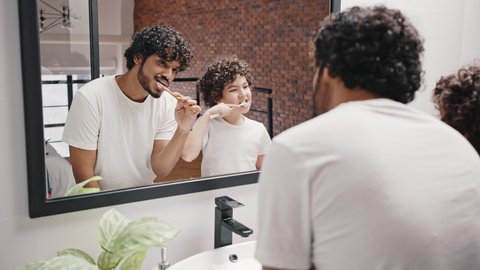 Asian Indian man and biracial brother brush teeth against mirror after waking up in morning. Curly-haired siblings keep routine in bathroom closeup