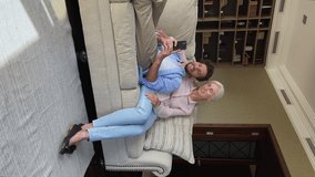 Vertical view millennial male lie on couch lean back to beloved senior mom grandma show picture video at social media to grand parent on phone. Loving grey haired old mum adult son relax using gadget
