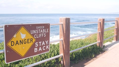 Ocean waves crashing on beach or bluff, La Jolla shore waterfront promenade, California USA. Succulent green ice plant, pacific coast. Seascape view and railings. Danger unstable cliff warning sign.