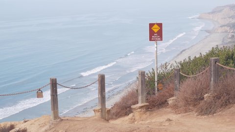 Steep unstable cliff, rock or bluff, foggy weather, California coast erosion, USA. Torrey Pines eroded crag overlook viewpoint, ocean waves from above. Chain railing for safety trekking, danger sign.