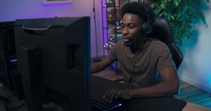 Pro male gamer is focused on winning round has headset talks to friends jokes, online friendships, backlit keyboard and mouse to play on computer, purple and blue neon lights in room.