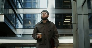 A young bearded man in a knitted hat drinks coffee from a disposable cup while walking near a stylish building with large windows, holding a laptop in a case in his hand. Video in 4k, red komodo