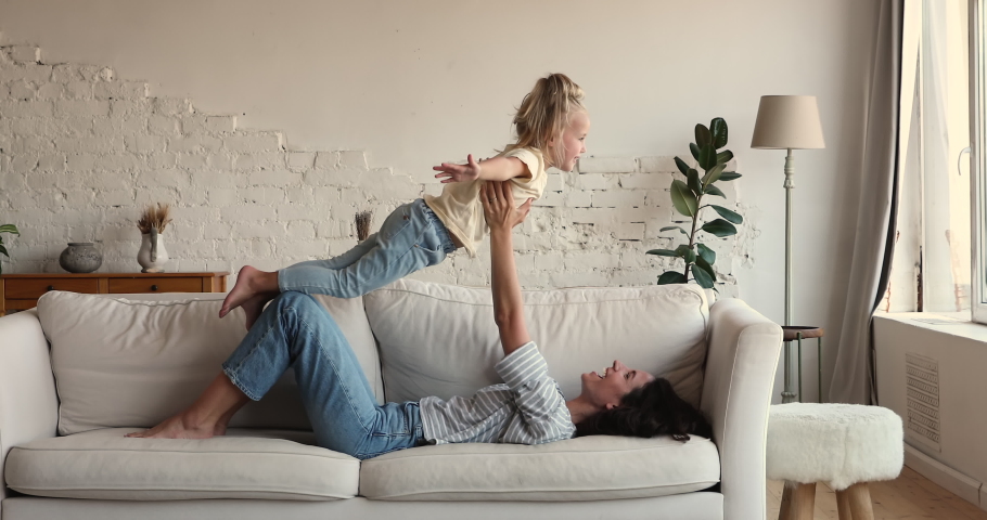 Happy caring mom play on sofa with little daughter lie on couch support girl in air on bent legs imitate acrobatic exercises. Sporty young mother child preschooler do gymnastics relax pretend flight Royalty-Free Stock Footage #1089354895