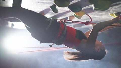 Female climber training on a climbing wall, young woman practicing rock-climbing and moving up, vertical video, rises using insurance, cinematic light.