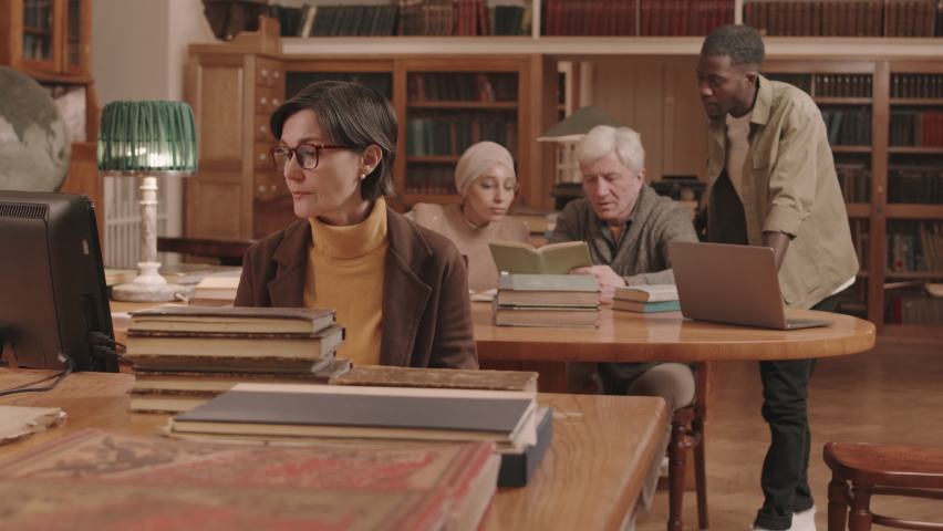 Medium slowmo of mature female librarian in eyeglasses sorting and classifying books at workplace while senior professor and his two diverse university students studying at desk in background