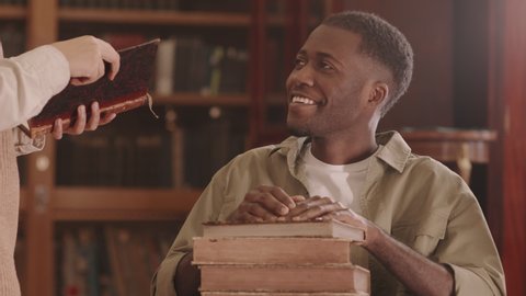 Tilt slowmo of smiling young Black man with disability sitting with pile of books on his laps in wheelchair talking to librarian at university library