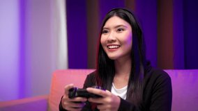 Playing video game. Young asian pretty woman sitting on sofa holding joystick in living room. Happy female Professional Streamer chinese playing game online in dark room neon light.