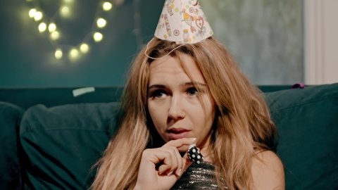 Close-up portrait of upset and sad lonely blonde girl lying or sitting on sofa or couch in birthday or festive cap and whistling whistle. Young woman in decorated house celebrating Party alone.