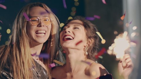 Close up couple of young multiracial girls wearing plush dresses enjoying glamorous party and having fun. Women dancing with falling confetti and sparkling or glowing sparklers or fireworks.