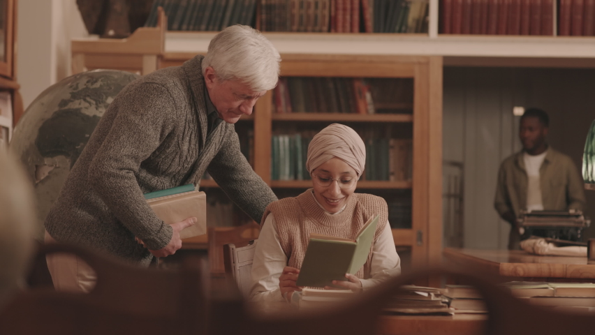 Medium slowmo of young Muslim woman in headscarf sitting by wooden desk at library with book in hand discussing plot with Caucasian senior man standing by her side
