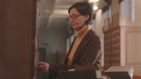 Medium side view of mature female librarian in eyeglasses sorting and classifying library card catalogue opening and closing drawers