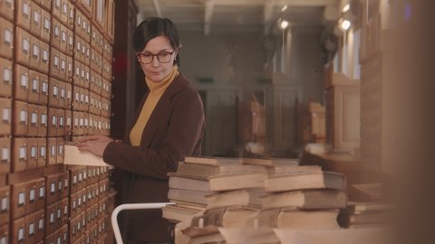 Medium slowmo of mature Caucasian female librarian in eyeglasses standing by drawers with library paper database or archive searching for necessary book