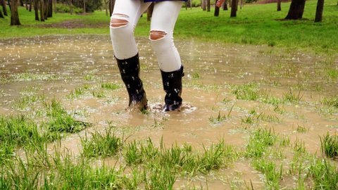Close-up of unrecognizable woman jumping with galoshes on pond. Horizontal low angle view of wellingtons on rainy pound splashing with mud. Nature and people backgrounds.