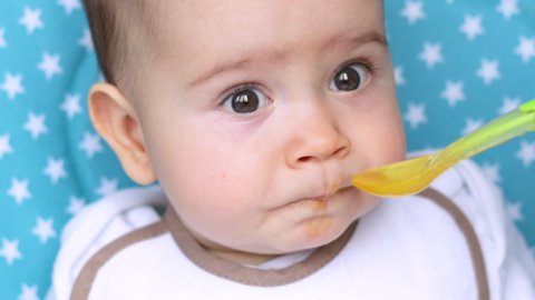 very close up video of a baby boy that is feed by mother or babysitter. cute toddler with beautiful big eyes. baby is eating mashed vegetables or fruit with green spoon. food diversification concept.
