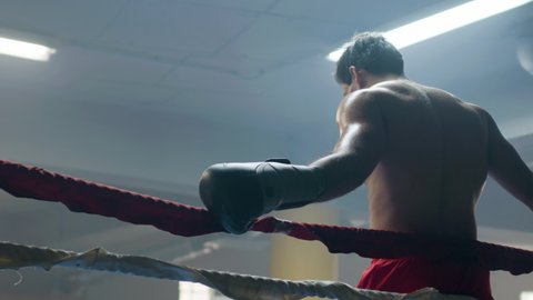 Young male boxer leaning on ring rope, then doing shadow fight while training in boxing gym alone. Athletic man practicing punches and dodging techniques. Sport, boxing concept