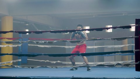Young male boxer doing shadow fight while training in boxing ring alone. Athletic man practicing punches, self-defense and dodging techniques. Sport, boxing concept