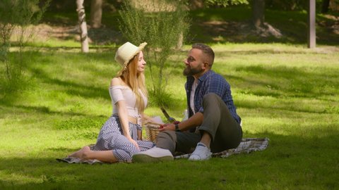 Romantic summer date. Outdoor portrait of happy loving couple enjoying picnic in green summer park, sitting on grass, eating strawberries and kissing, slow motion