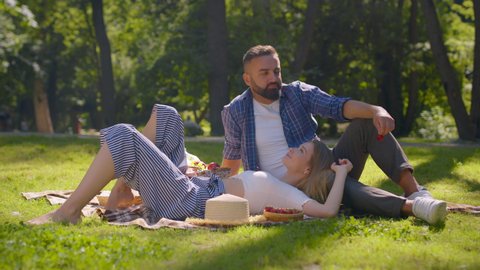 Summer romance. Happy couple in love enjoying picnic in public park, man feeding woman with ripe strawberries, resting on green grass, slow motion