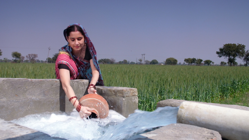A married Indian housewife in traditional Sari filling up fresh water for daily use - cooking, an Indian villager. A female worker laborer filling up an earthen pot from a tubewell near a rice field.. Royalty-Free Stock Footage #1089368135