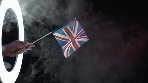 Union Jack. United Kingdom. Great Britain. Patriotic symbol. Female hand waving British flag with red cross on white blue in smoke cloud LED light ring isolated on black.