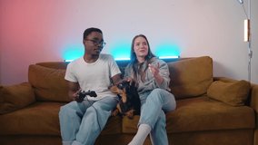 A cute dachshund is lying on a couch between a mixed young couple who are playing video games and laughing. Slow motion