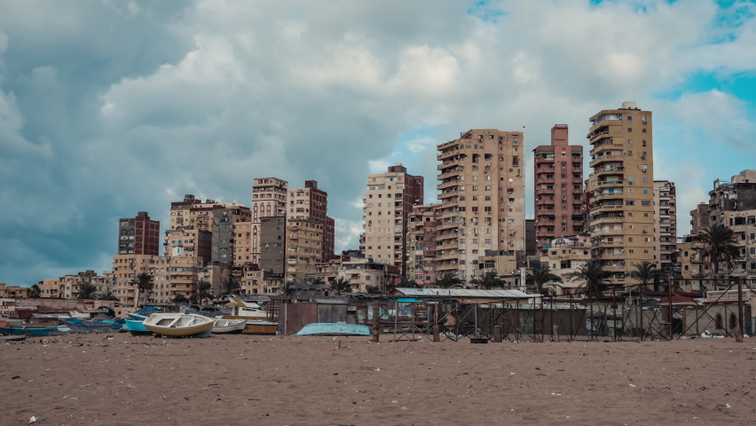 Sea coast of Alexandria. Seashore of ancient city of Egypt with buildings, constructions, palms, boats. Timelapse. Urban view of seaside with houses, moving people, trees, ships. Cloudy day