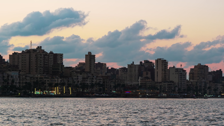 Sea coast of Alexandria. Seashore of ancient city of Egypt with buildings, constructions, palms, boats. Timelapse. Evening sunset time. Urban view of seaside with houses, ships. Cloudy day