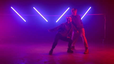 Professional dancers performing street style on nightclub stage. Active guy dancing behind twerking girl illuminated ultraviolet lights. Attractive couple moving body in freestyle standing scene.