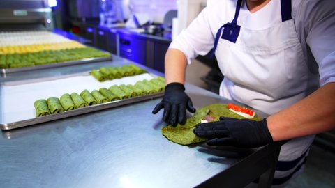 Female restaurant cook covers the stuffing in a green crepe. Ready rolled pancakes are beautifully laid on the metal trays. Blurred backdrop.
